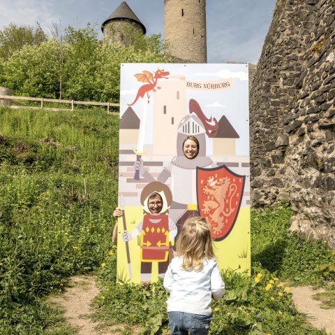 Ein Kind betrachtet einen Fotostand mit Rittermotiven vor der Burgruine Nürburg. Im Hintergrund sind die Türme der Burg zu sehen., © Eifel Tourismus GmbH,D.Ketz Ein Kind betrachtet einen Fotostand mit Rittermotiven vor der Burgruine Nürburg. Im Hintergrund sind die Türme der Burg zu sehen., © Eifel Tourismus GmbH,D.Ketz