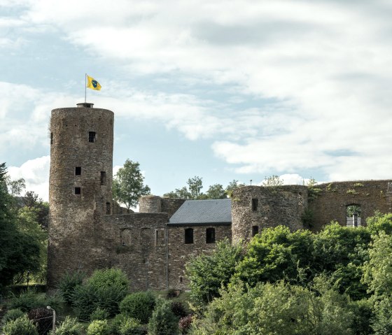 Burg Reuland mit einem runden Turm und einer gelben Flagge, umgeben von grünen Bäumen und bewölktem Himmel., © vennbahn.eu Burg Reuland mit einem runden Turm und einer gelben Flagge, umgeben von grünen Bäumen und bewölktem Himmel., © vennbahn.eu