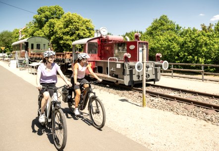 Zwei Radfahrerinnen auf einem Radweg neben einer alten Lokomotive im Eisenbahnmuseum Pronsfeld, umgeben von grünen Bäumen., © Eifel Tourismus GmbH, Dominik Ketz Zwei Radfahrerinnen auf einem Radweg neben einer alten Lokomotive im Eisenbahnmuseum Pronsfeld, umgeben von grünen Bäumen., © Eifel Tourismus GmbH, Dominik Ketz