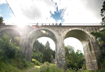 Radfahrer überqueren das Reichensteiner Viadukt der Vennbahn. Die Sonne scheint durch die Wolken, während das Viadukt über eine grüne Landschaft führt., © vennbahn.eu Radfahrer überqueren das Reichensteiner Viadukt der Vennbahn. Die Sonne scheint durch die Wolken, während das Viadukt über eine grüne Landschaft führt., © vennbahn.eu