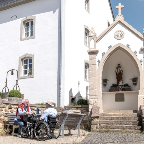 Zwei Männer, einer im Rollstuhl, sitzen auf einer Bank vor einem weißen Gebäude mit einer religiösen Statue in einer Nische. Sonniger Tag., © Naturpark Südeifel, Thomas Urbany Zwei Männer, einer im Rollstuhl, sitzen auf einer Bank vor einem weißen Gebäude mit einer religiösen Statue in einer Nische. Sonniger Tag., © Naturpark Südeifel, Thomas Urbany
