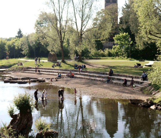 Menschen entspannen im Kyllpark am Wasser, während ein Kirchturm im Hintergrund zu sehen ist. Kinder spielen im flachen Wasser., © Clara Zins-Grohé Menschen entspannen im Kyllpark am Wasser, während ein Kirchturm im Hintergrund zu sehen ist. Kinder spielen im flachen Wasser., © Clara Zins-Grohé