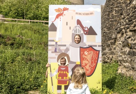 A child looks at a photo stand with knight motifs in front of the ruins of Nürburg Castle. The towers of the castle can be seen in the background., © Eifel Tourismus GmbH,D.Ketz A child looks at a photo stand with knight motifs in front of the ruins of Nürburg Castle. The towers of the castle can be seen in the background., © Eifel Tourismus GmbH,D.Ketz