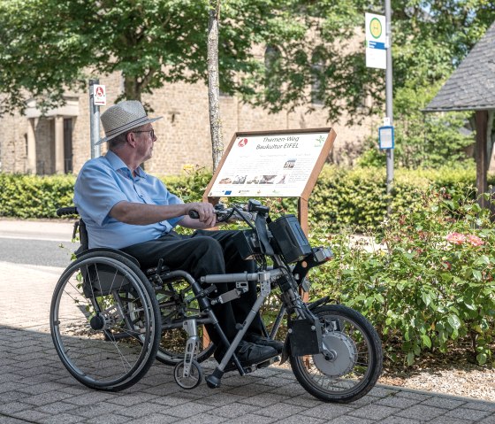 A man in a wheelchair reads a sign for the Eifel building culture theme trail in Wolsfeld. Trees and a building can be seen in the background., © Naturpark Südeifel, Thomas Urbany A man in a wheelchair reads a sign for the Eifel building culture theme trail in Wolsfeld. Trees and a building can be seen in the background., © Naturpark Südeifel, Thomas Urbany