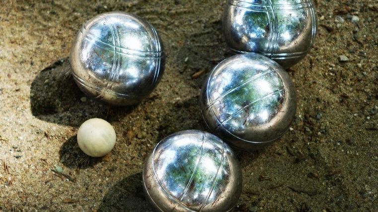 Un ensemble de quatre boules de pétanque en métal est posé dans le sable, à côté d'une petite boule blanche. L'environnement est naturel et ensoleillé.
