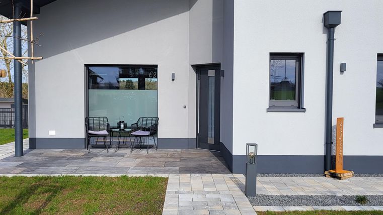 Ein modernes Haus mit einem grauen Anstrich und großen Fenstern. Der Weg zur Eingangstür ist mit Pflastersteinen gestaltet und führt über eine grüne Rasenfläche.