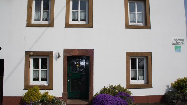 Ein charmantes, zweigeschossiges Haus mit weißen Wänden und braunen Fensterrahmen. Vor dem Eingang blühen bunte Pflanzen.