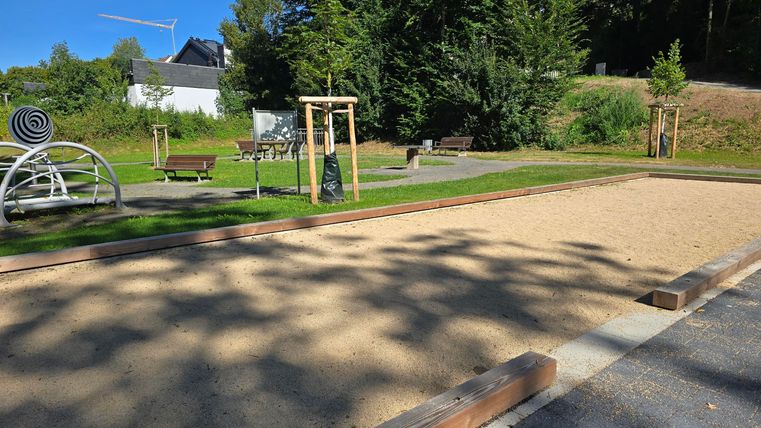 Ein kinderfreundlicher Spielplatz mit Sandfläche und Spielgeräten. Im Hintergrund sind grüne Bäume und Sitzgelegenheiten zu sehen.