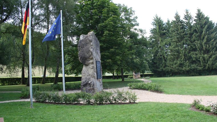 Ein Denkmal in einem Park, umgeben von Bäumen und Sträuchern. Zwei Flaggen wehen daneben, eine deutsche und eine europäische.