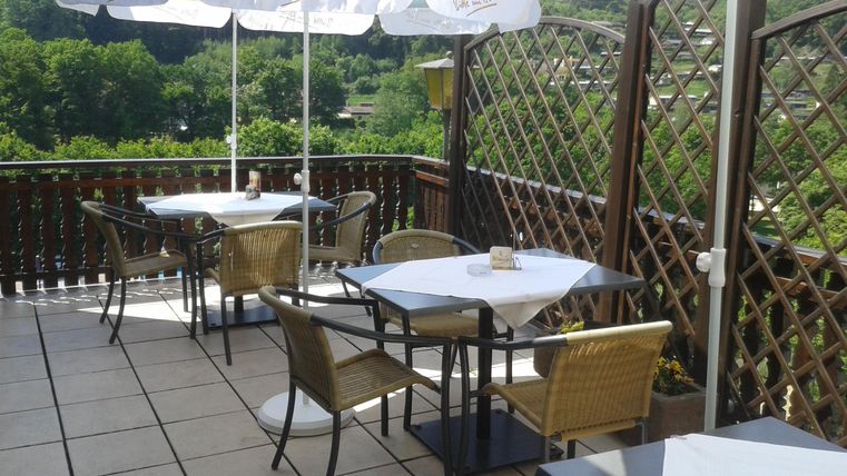 Eine Terrasse mit Tischen und Stühlen, umgeben von einem Gitterzaun. Sonnenschirme bieten Schatten und die Umgebung besteht aus grünen Bäumen.