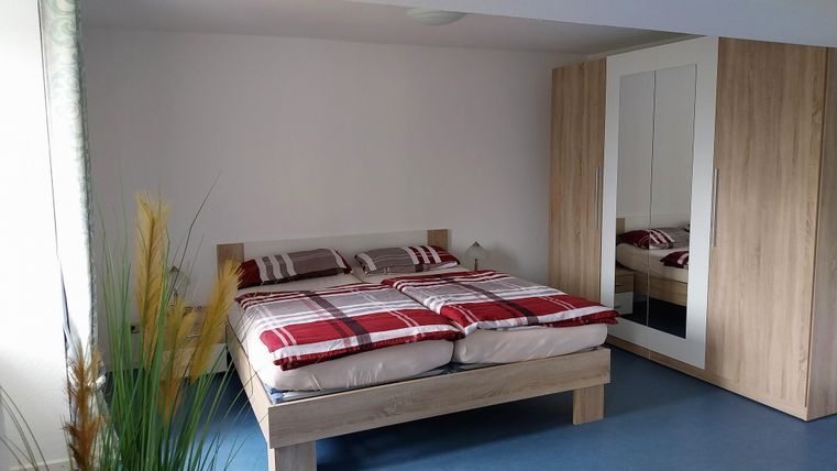 Ein modernes Schlafzimmer mit einem großen Bett und karierter Bettwäsche. Ein schmaler Kleiderschrank und grüne Pflanzen sorgen für eine gemütliche Atmosphäre.