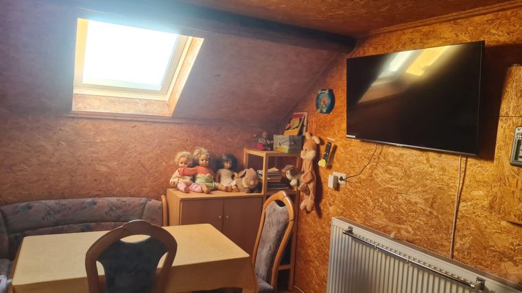 Ein gemütlicher Raum mit einem Tisch und Stühlen. Ein Dachfenster lässt Tageslicht herein, und an der Wand hängt ein Fernseher.