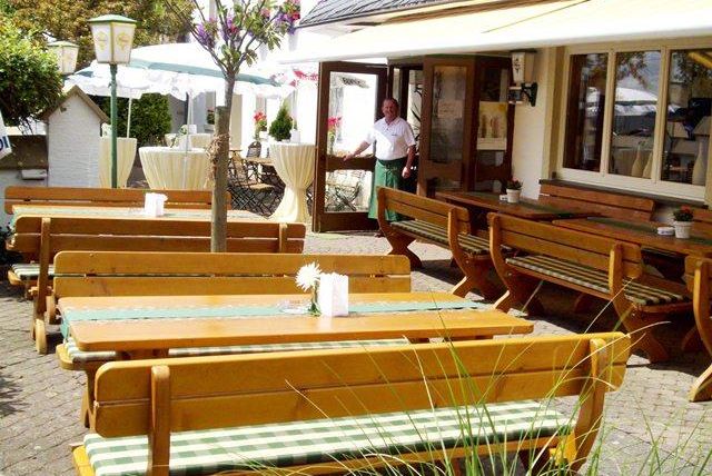 Ein einladender Außenbereich eines Restaurants mit Holztischen und Bänken. Im Hintergrund ist eine Person zu sehen, die das Restaurant betritt.