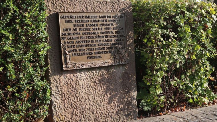 A commemorative plaque made of stone with an inscription in memory of historical events. It stands in a planted area next to a walkway.