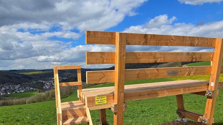 Eine Holzbänke mit Treppe steht auf einem Hügel mit Blick auf eine Landschaft. Der Himmel ist teilweise bewölkt und das Grün der Wiesen ist gut sichtbar.