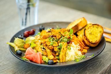Ein bunter Teller mit verschiedenen Salaten, Gemüse und Hähnchenstücken. Dazu gibt es knuspriges Brot und frisches Obst.
