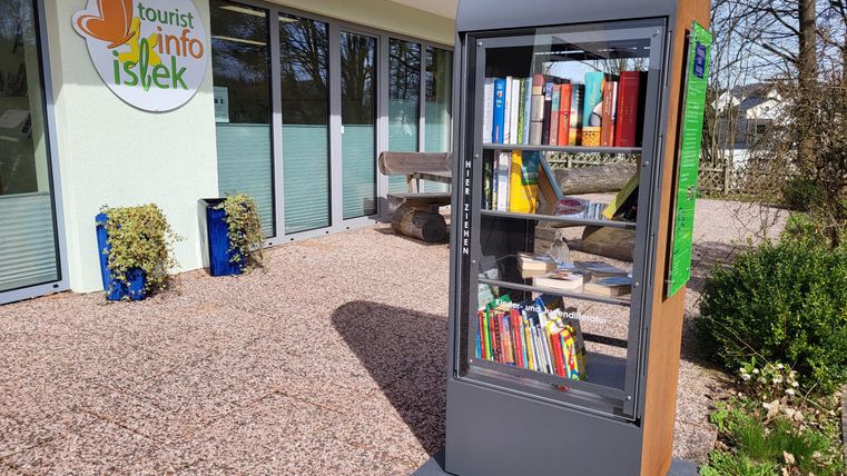 Ein Bücherregal steht vor einem Informationszentrum. Es ist mit verschiedenen Büchern gefüllt und befindet sich in einer begrünten Umgebung.