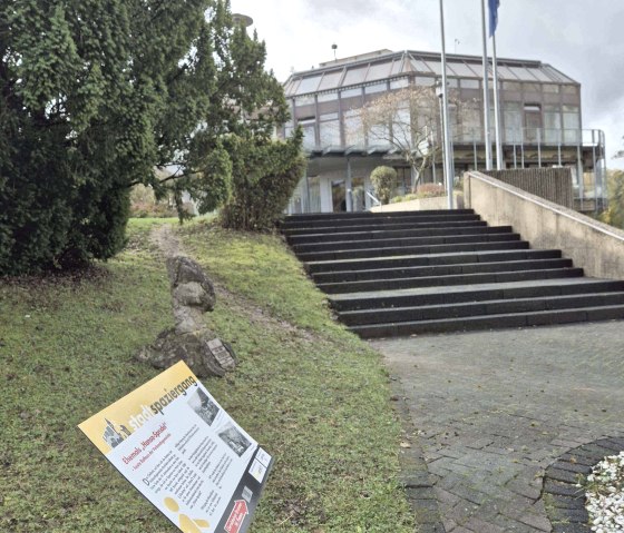 Modernes Geb&auml;ude mit Glasfassade, Treppe und Flaggen. Im Vordergrund ein Schild auf einer Wiese, umgeben von B&auml;umen., &copy; Clara Zins-Groh&eacute;