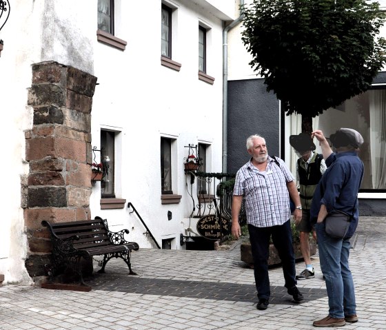 Zwei M&auml;nner stehen vor einem alten Torbogen. Einer zeigt gestikulierend, w&auml;hrend der andere zuh&ouml;rt. Im Hintergrund ein Baum und ein Geb&auml;ude., &copy; Clara Zins-Groh&eacute;