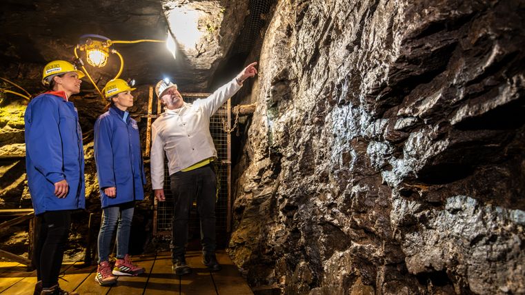 Drei Personen in Schutzkleidung im Bergwerk, eine Person zeigt auf die Wand.