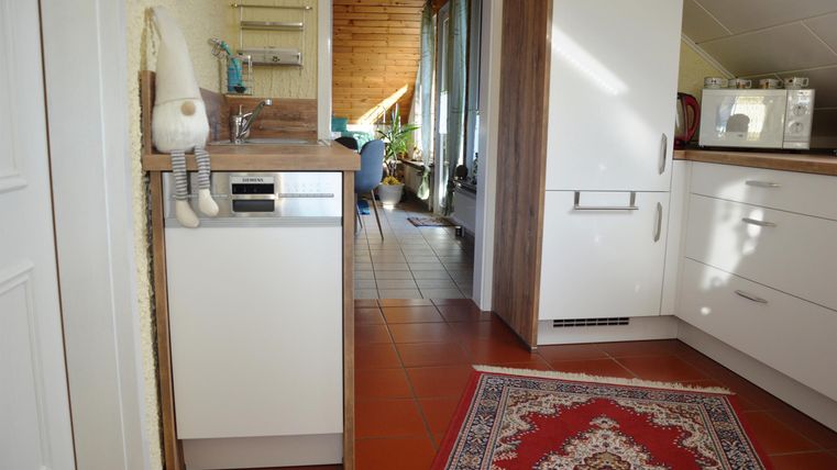 Eine moderne Küche mit weißen Schränken und einem Teppich im Vordergrund. Im Hintergrund sind Fenster und einige Möbel sichtbar.