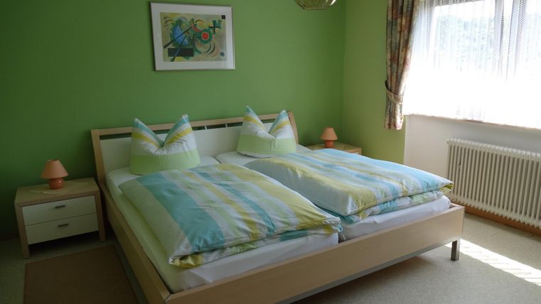 A modernly furnished bedroom with a large double bed and green walls. The room has a window with plenty of daylight and stylish lighting.