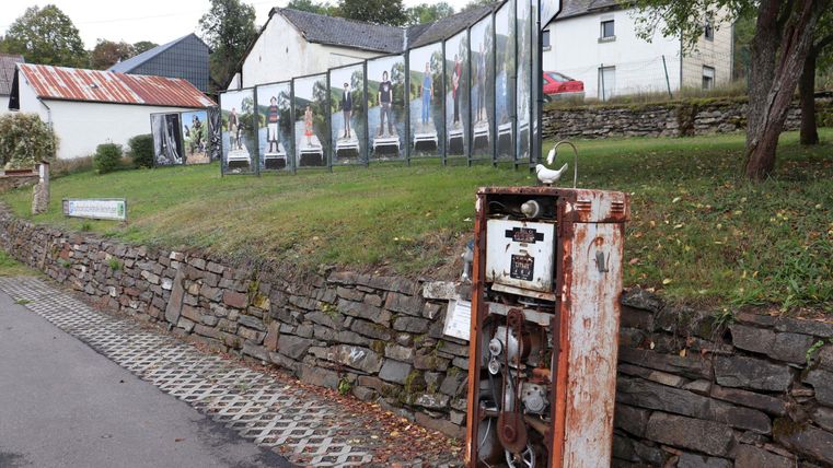 Une vieille pompe à essence rouillée se trouve contre un mur en pierre. En arrière-plan, des photos dans de grands cadres sont exposées, montrant une exposition.