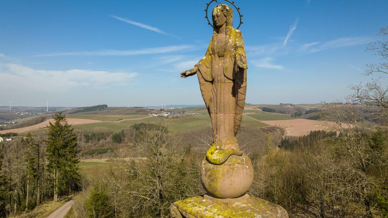 Eine Statue der Jungfrau Maria steht auf einem hohen Sockel und blickt über die umliegende Landschaft. Der Himmel ist klar und es gibt grüne Hügel und einen Kiesweg im Vordergrund.
