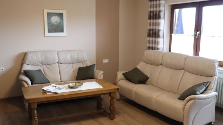 Ein gemütliches Wohnzimmer mit zwei beigen Sofas und einem Holzkaffee Tisch. An der Wand hängt ein Bild und das Licht scheint durch die Fenster.