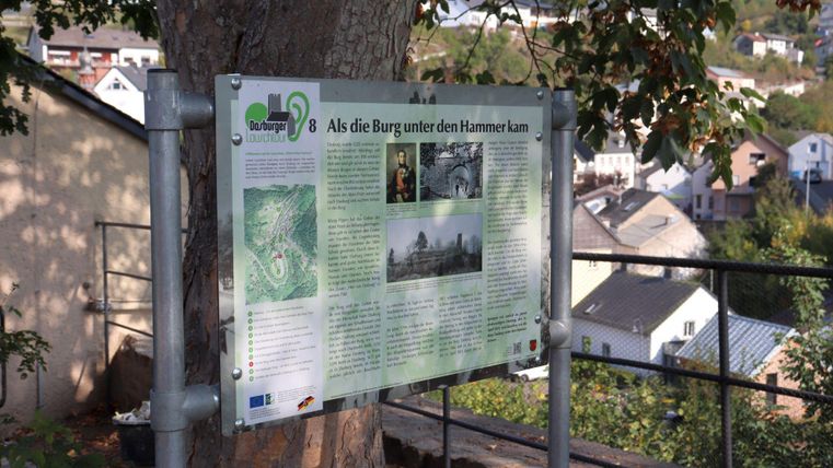 Ein Informationstafel an einem Baum, die über die Burg und ihre Geschichte informiert. Im Hintergrund sind Häuser und Bäume sichtbar.