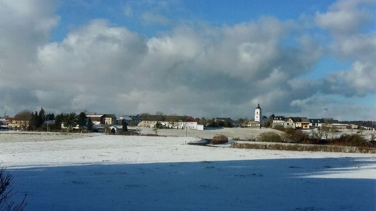 Eine winterliche Landschaft mit schneebedecktem Feld und einem kleinen Dorf im Hintergrund. Der Himmel ist teilweise bewölkt und strahlt in verschiedenen Blautönen.