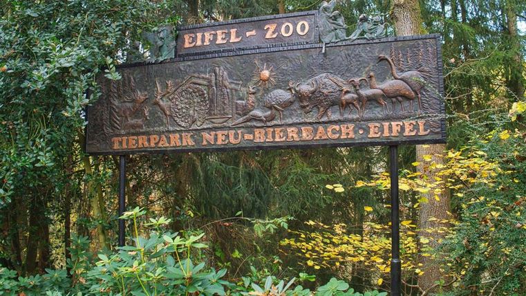 Ein gut gestaltetes Schild des Eifel-Zoo, umgeben von Bäumen und Pflanzen. Der Zoo lädt Besucher ein, die Tierwelt zu entdecken.