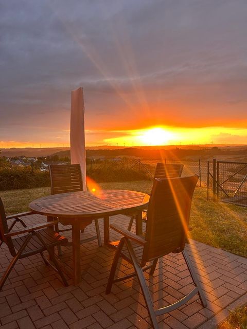 Eine Terrasse mit einem Tisch und Stühlen, die bei Sonnenuntergang fotografiert wurde. Die warmen Farben des Sonnenuntergangs erhellen die Landschaft im Hintergrund.