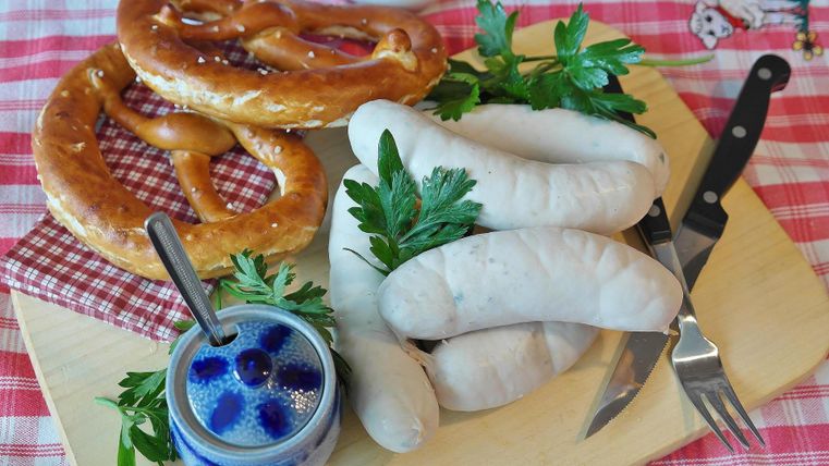 Ein Teller mit bayerischen Spezialitäten: Weisswurst, Brezeln und süßer Senf, garniert mit frischer Petersilie. Dazu Besteck und ein kariertes Tuch auf einem Holzbrett.