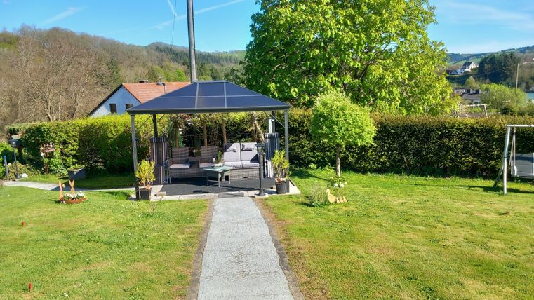Eine gemütliche Gartenlandschaft mit einem Pavillon und viel Grün. Der Weg führt zum Pavillon, umgeben von Pflanzen und Bäumen.