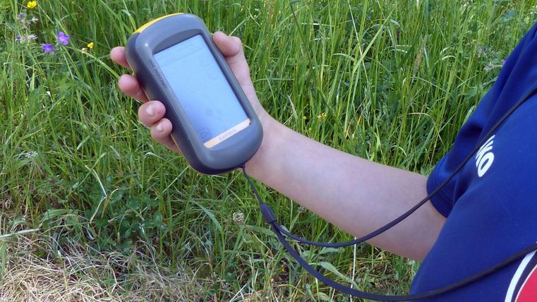 Ein Mensch hält ein Handgerät in der Hand, während er in einem grünen, mit Gras und Blumen bewachsenen Gebiet steht. Das Gerät scheint für Navigation oder Messungen verwendet zu werden.