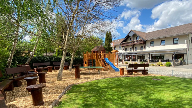 Ein schöner Außenbereich mit einer Spielecke, Holzsitzen und einer Rutsche. Im Hintergrund befindet sich ein gemütliches Gebäude mit Terrasse.