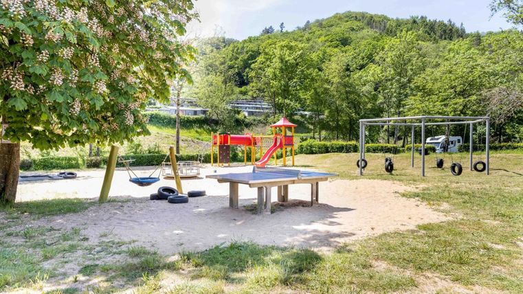 Ein Spielplatz mit Schaukeln, einer Rutsche und einer Tisch-Anlage. Umgeben von grünen Bäumen und Wiesen.