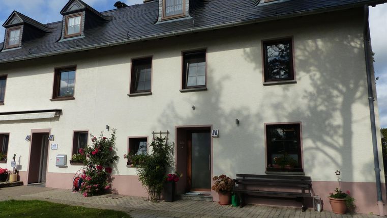 Eine schöne, zweigeschossige Wohnung mit einem hellen Außenbereich. Vor dem Haus stehen Blumen und eine Bank lädt zum Verweilen ein.