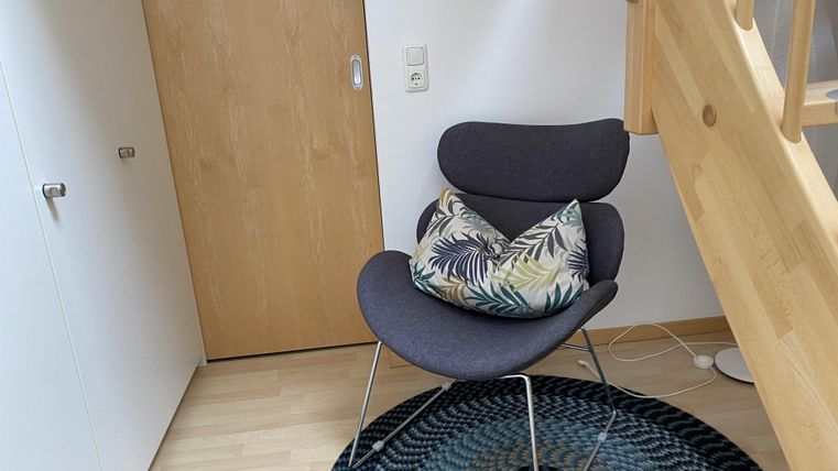 Een comfortabele stoel met een kleurrijk kussen staat in een hoek van een lichte kamer. De vloer is bedekt met een rond tapijt, en een houten trap leidt naar boven.