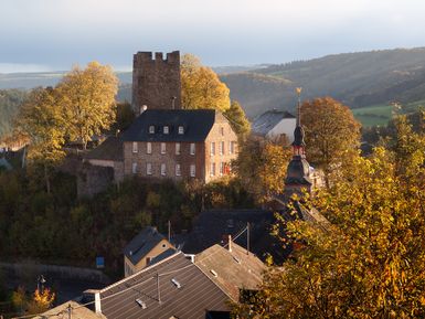 Eine malerische Landschaft mit einer alten Burg und bunten Bäumen im Herbst. Im Vordergrund sind charmante Dächer von Häusern sichtbar.
