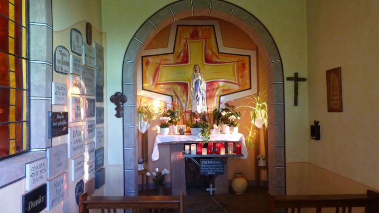 Eine kleine Kapelle mit einem Altar, umgeben von Kerzen und Pflanzen. An der Wand sind Bilder und eine große Kreuzdarstellung zu sehen.
