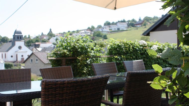 Eine gemütliche Terrasse mit einem Tisch und Stühlen aus Rattan. Im Hintergrund sieht man eine malerische Landschaft mit Häusern und einem Kirchturm.