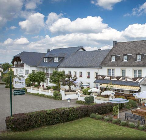Ein einladendes Hotel mit einer schönen Terrasse und vielen Sitzplätzen im Freien. Der Himmel ist blau mit einigen Wolken und die Umgebung ist gepflegt.