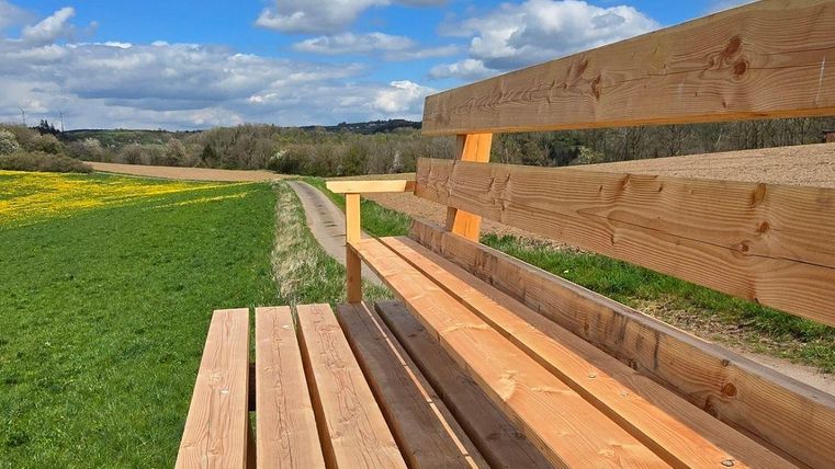 Eine Holzbank steht auf einem grünen Feld unter einem blauen Himmel mit Wolken. Im Hintergrund sind Wiesen und Bäume zu sehen.