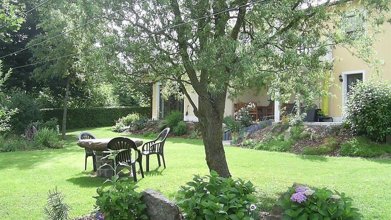 Un beau jardin avec une pelouse verte et un grand arbre. On y voit une table et des chaises sous l'arbre ainsi que des parterres de fleurs colorées.
