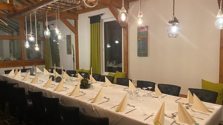 Ein eleganter Essbereich mit einem langen Tisch, der festlich gedeckt ist. Warmes Licht von hängenden Lampen schafft eine einladende Atmosphäre.