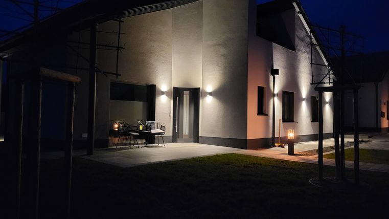 Ein modernes Haus bei Nacht, sanft beleuchtet von Außenlichtern. Vor dem Eingang stehen Stühle und ein Tisch, umgeben von einem ruhigen Garten.