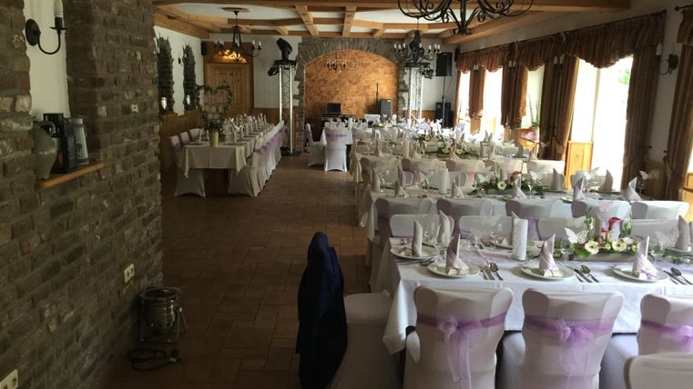 Ein festlich dekorierter Saal mit langen Tischen. Die Tische sind mit weißen Tüchern und lila Schleifen geschmückt, bereit für eine Feier.