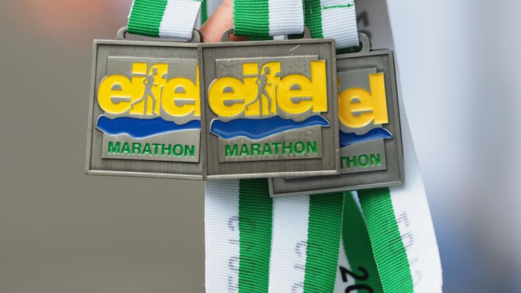 Drei Medaillen für den Eifel-Marathon hängen an grünen und weißen Bändern. Die Medaillen sind aus Metall und tragen das Logo des Marathons.
